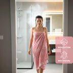 Woman wearing a pink floral towel in a bathroom with a '100% Cotton' and 'Skin Friendly' label. VU-BLO-PNK